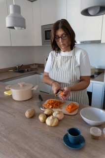 Woman wearing apron preparing vegetables for homemade soup while peeling carrots in modern kitchen.