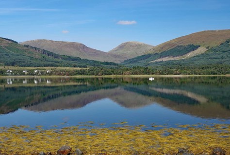 A calm body of water reflects the surrounding mountains and clear skies, Highlands, Fort William,