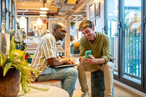 Two diverse male colleagues are sharing information on a mobile phone and laptop while working