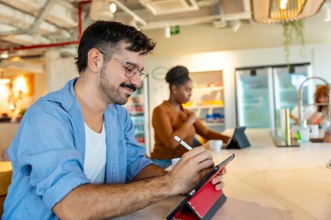 Man in casual attire smiling while using a digital tablet with a stylus to sketch ideas at a bar