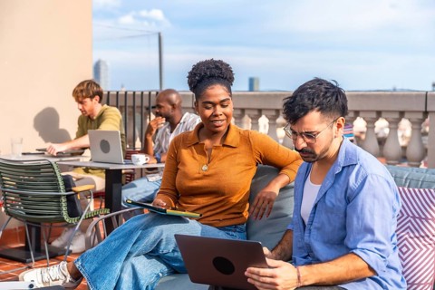 Diverse colleagues working and interacting on a rooftop terrace, using laptops and tablets while