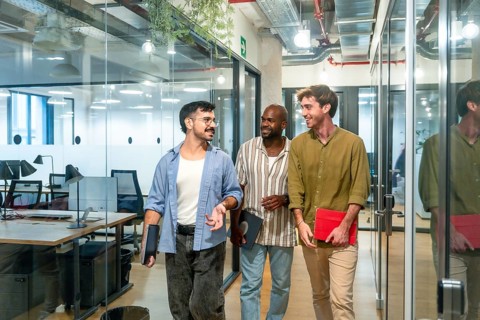 Three diverse male colleagues walking through a modern office corridor, smiling and engaging in