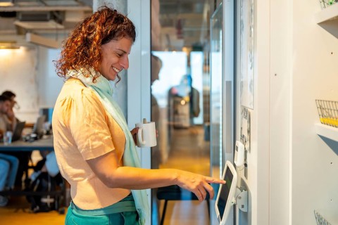 Woman standing in a modern office or coworking space, smiling and holding a coffee cup while using