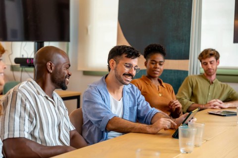 Diverse group of smiling colleagues interacting and working together at a conference table, using a