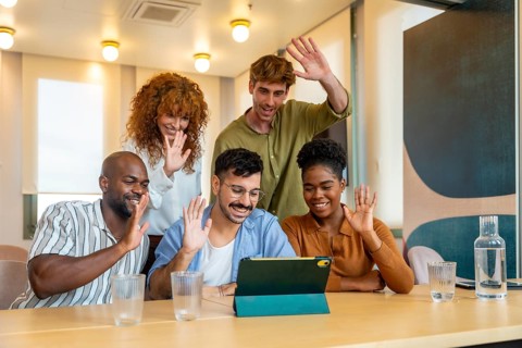 Group of diverse colleagues waving to a tablet during a business video conference in a modern