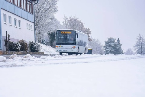 A bus travels on a snowy road surrounded by trees and buildings, Gechingen, Calw district, Germany