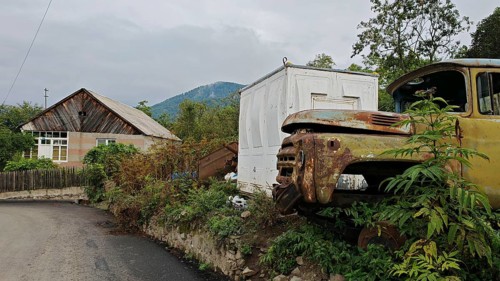 A rusty truck is parked in front of a small house on the side of the road in a village, retro