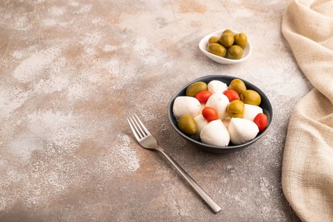 White Mozzarella cheese, with tomatoes and olives in blue bowl on brown concrete background and