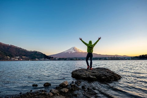 Young man stretching his arms in the air, standing on rocks in Lake Kawaguchi, view of Mount Fuji