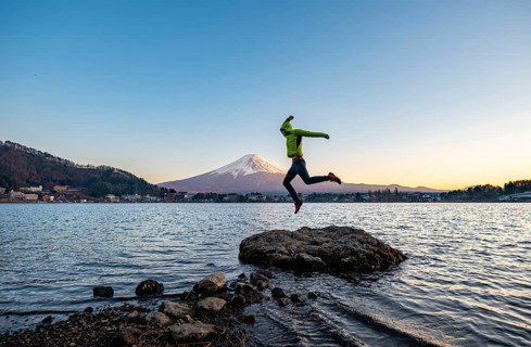Young man jumping on rocks in Lake Kawaguchi, view of Mount Fuji volcano at sunset, Yamanashi