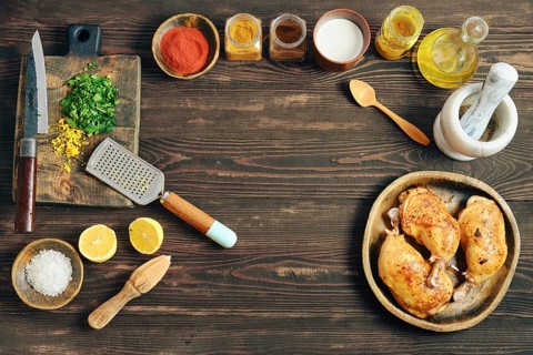 Roasted chicken legs are on a wooden platter next to bowls of spices, herbs, and lemon. A knife,