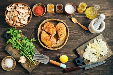 Top view of fried chicken thighs on a wooden plate. Chopped onions, mushrooms, and herbs are placed
