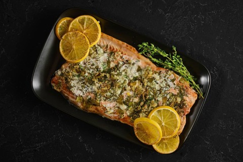 Roasted pink salmon is served on a black plate with slices of lemon and herbs. This dish is ready