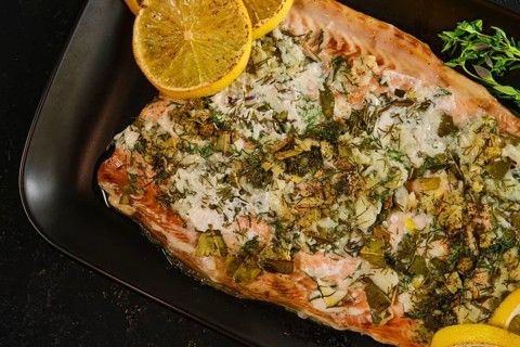 A pink salmon fillet is ready for serving on a black plate. It is garnished with herbs and slices