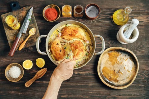 A hand puts fried chicken thighs in a saucepan with creamy mushroom sauce, top view. The pan is