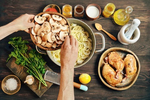 Two hands hold a bowl of sliced mushrooms over a pot with cooking onions on a wooden table.