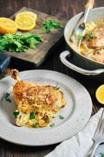 A chicken portion on a plate, surrounded by lemon slices and herbs. Cooking pan with more chicken