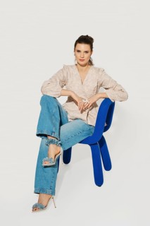 A woman sits on a blue chair with one leg crossed over the other. She wears a light top and denim