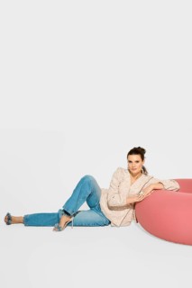 A woman sits on a floor near a pink piece of furniture in a bright studio. She leans back, wearing
