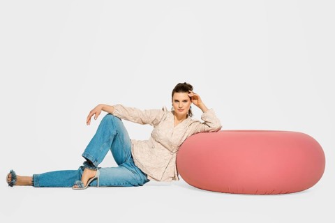 Cute woman is sitting on the floor beside a round pink chair in a studio. She is wearing a light