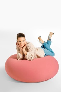 A woman lays on a big pink cushion in a studio. She is wearing a light blazer and jeans. Her hair