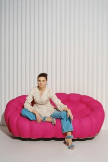 Woman sits with one leg tucked under her on a pink couch in a studio. She wears a light blazer and