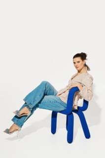A model sits on a blue chair, leaning back with one leg crossed over the other. She wears a light