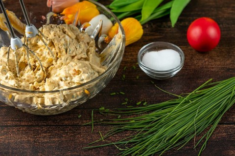 Egg salad in a glass bowl with a mixer, fresh ingredients in the background