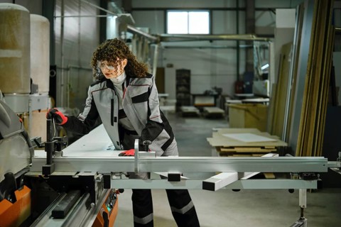 Woman is focused on using a large circular saw in a factory. The worker wears safety gear and