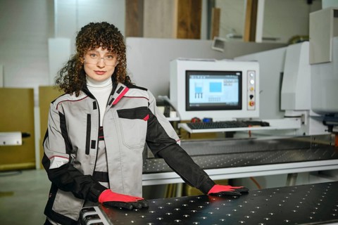 Portrait of female worker next to a large cutting machine in a furniture workshop. She wears gloves