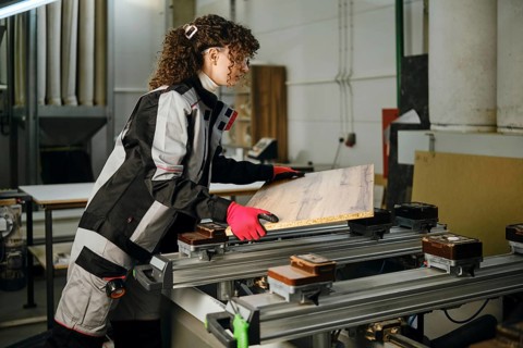 A woman is in carpentry workshop handling a piece of wood. She wears safety gloves and works with