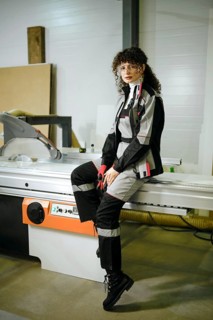 A woman is sitting on a large cutting machine in a workshop. She is dressed in work clothes and is