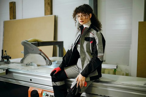 A woman sits on a machine in a workshop. She is wearing a work outfit and gloves. The workshop has