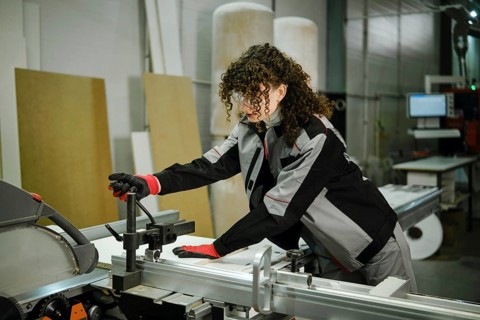 A woman in a workshop operates a table saw machine. She uses her hands to adjust a piece of wood.