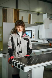 A woman stands in a workshop next to a wood shaping machine. She wears a work suit and gloves while