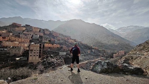 A hiker with backpack stands in front of a mountainous village in sunny weather, Atlas Mountains,