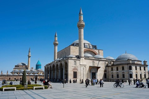 Historic mosque with several minarets under clear blue sky, people gathered in the square, Mevlânâ