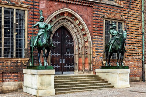 Two equestrian statues in front of a Gothic building with decorated entrance doors and red bricks,