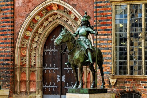 An equestrian statue in front of a Gothic building with richly decorated archway and red bricks,