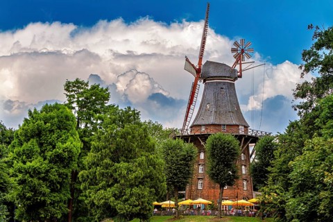 Historic windmill surrounded by trees under blue sky with white clouds and umbrellas, An old