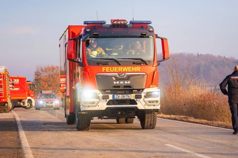 A fire truck drives on a road through a wooded area with emergency personnel, fire brigade exercise