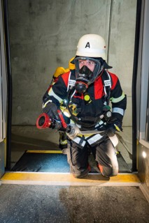 A firefighter wearing respiratory protection kneels in a tunnel at the threshold of a door, fire
