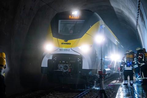 A train goes through an illuminated tunnel with firefighters in the background, fire department