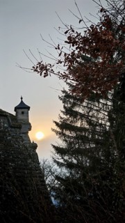 A castle tower, bay window of a fortress wall rising into the sky, surrounded by trees at sunset,