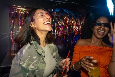 Two diverse young women enjoying a night out, laughing and dancing with drinks in hand amidst