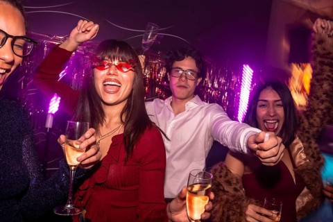 Young diverse friends dancing and cheering while holding glasses of alcoholic drinks, enjoying a