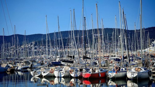 Numerous sailboats in harbor with reflecting masts in calm water against mountain backdrop,