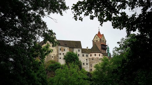 A historic castle rises behind thick greenery under a cloudy sky, Loket, Czech Republic
