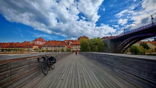 Wooden walkway with bicycle leading to a picturesque old town across the river under a blue sky
