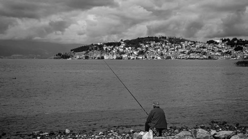 Black and white photo of a fisherman on a lake shore with a city in the background, Ohrid, North